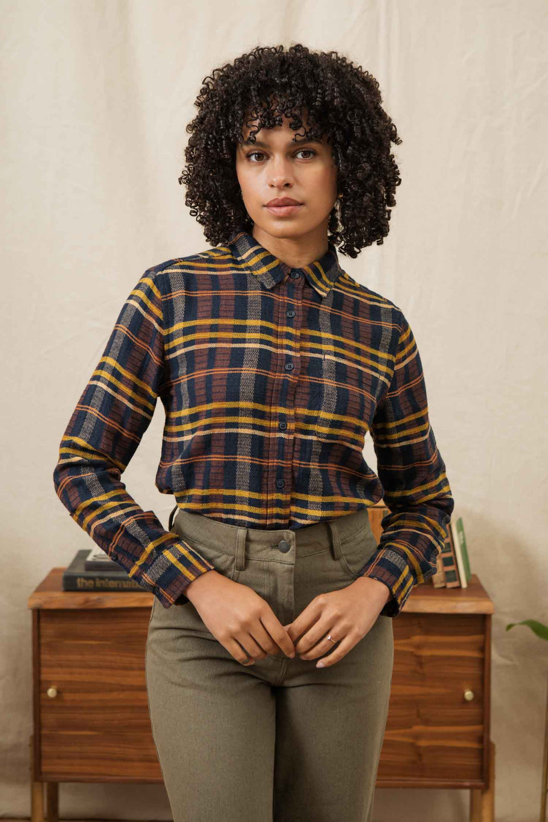 A person with curly hair wears the Bridge & Burn Ash Slim Shirt in Desert Dawn Plaid and high-waisted pants, standing before a wooden table with books and a plant, set against a light-colored background.