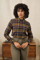 A person with curly hair wears the Bridge & Burn Ash Slim Shirt in Desert Dawn Plaid and high-waisted pants, standing before a wooden table with books and a plant, set against a light-colored background.