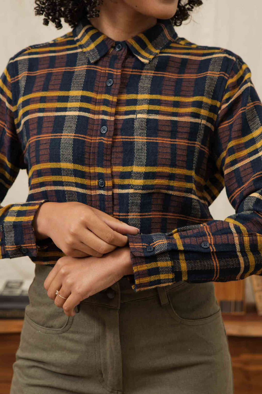 A person wears the Bridge & Burn Ash Slim Shirt in Desert Dawn Plaid—navy, yellow, and brown tones—with high-waisted olive green pants. Their hands are clasped at their waist.