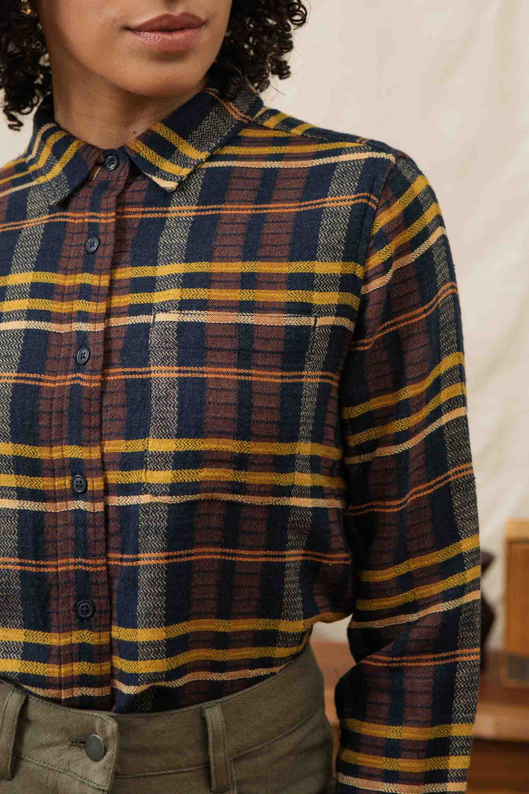 A person wearing the Bridge & Burn Ash Slim Shirt in Desert Dawn Plaid, featuring navy, yellow, and brown tones, paired with high-waisted olive green pants; only their torso and lower face are visible.