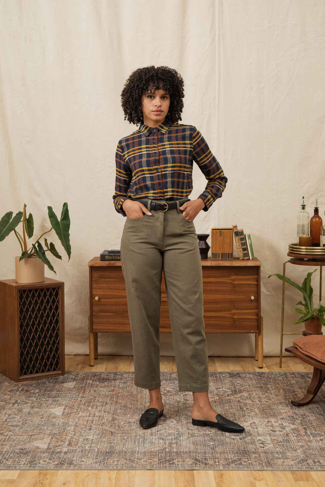 A woman with curly hair stands indoors on a patterned rug, wearing the Bridge & Burn Ash Slim Shirt in Desert Dawn Plaid, tucked into high-waisted olive pants with black loafers. Plants, a wooden sideboard, books, and bottles are in the background.