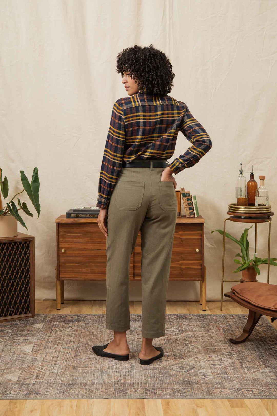 A person with curly hair stands with their back to the camera, wearing the Bridge & Burn Ash Slim Shirt in Desert Dawn Plaid, olive green pants, and black flats in a mid-century room with plants and wooden furniture.