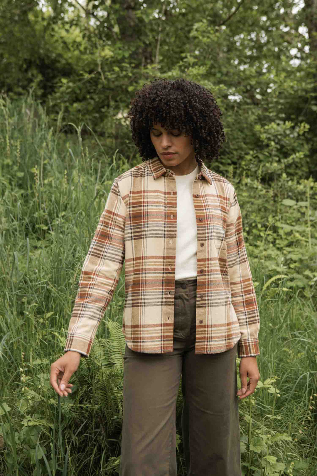 Amid tall grass and lush trees, a person with curly hair wears an off-white shirt and the Bridge & Burn Ash Slim Shirt in Latte Plaid, paired with olive green pants.