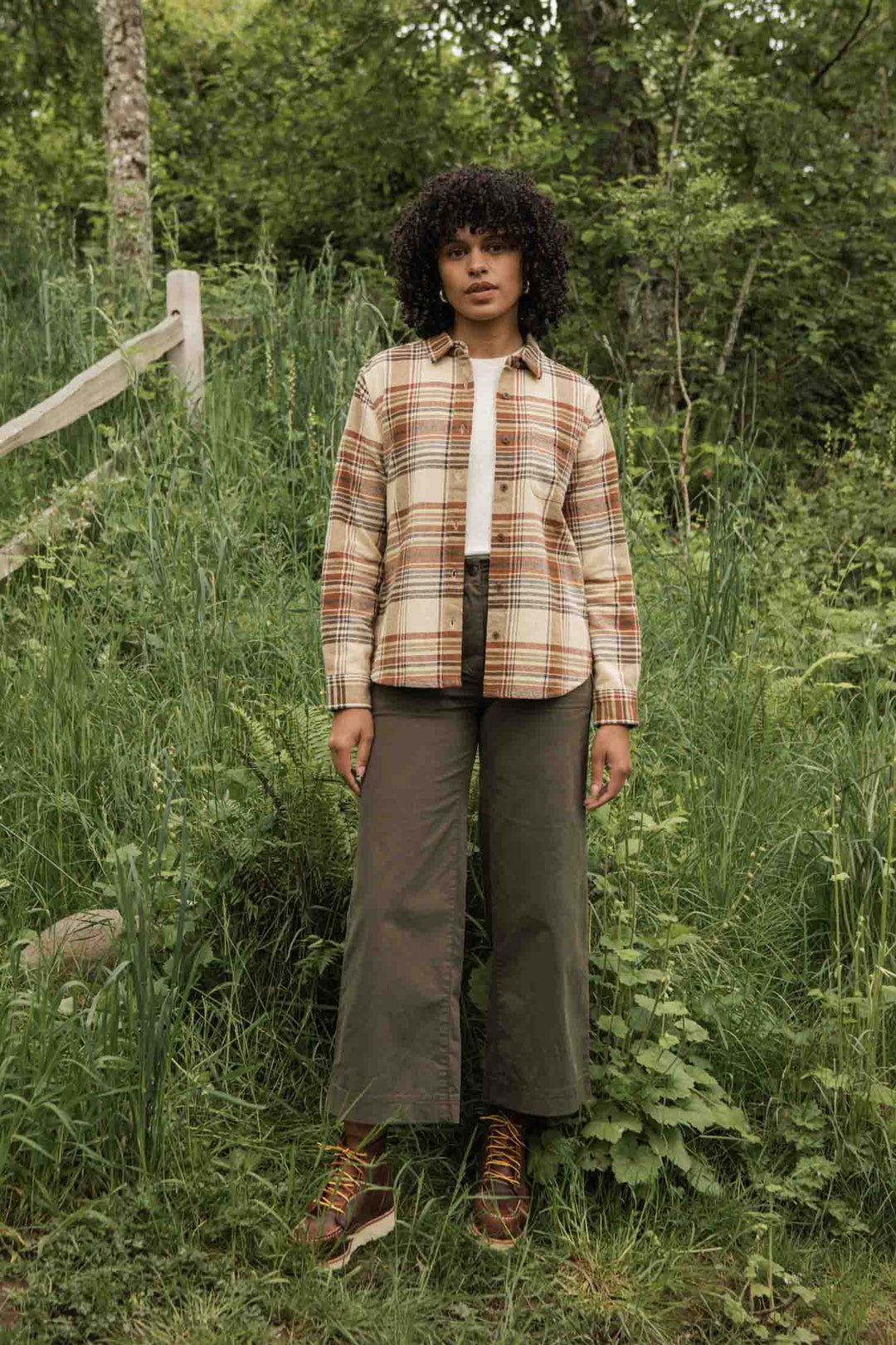 Standing outdoors in a grassy, wooded area, a person wears the Bridge & Burn Ash Slim Shirt in Latte Plaid over a white top, paired with green wide-leg pants and brown boots. A wooden fence appears in the background.
