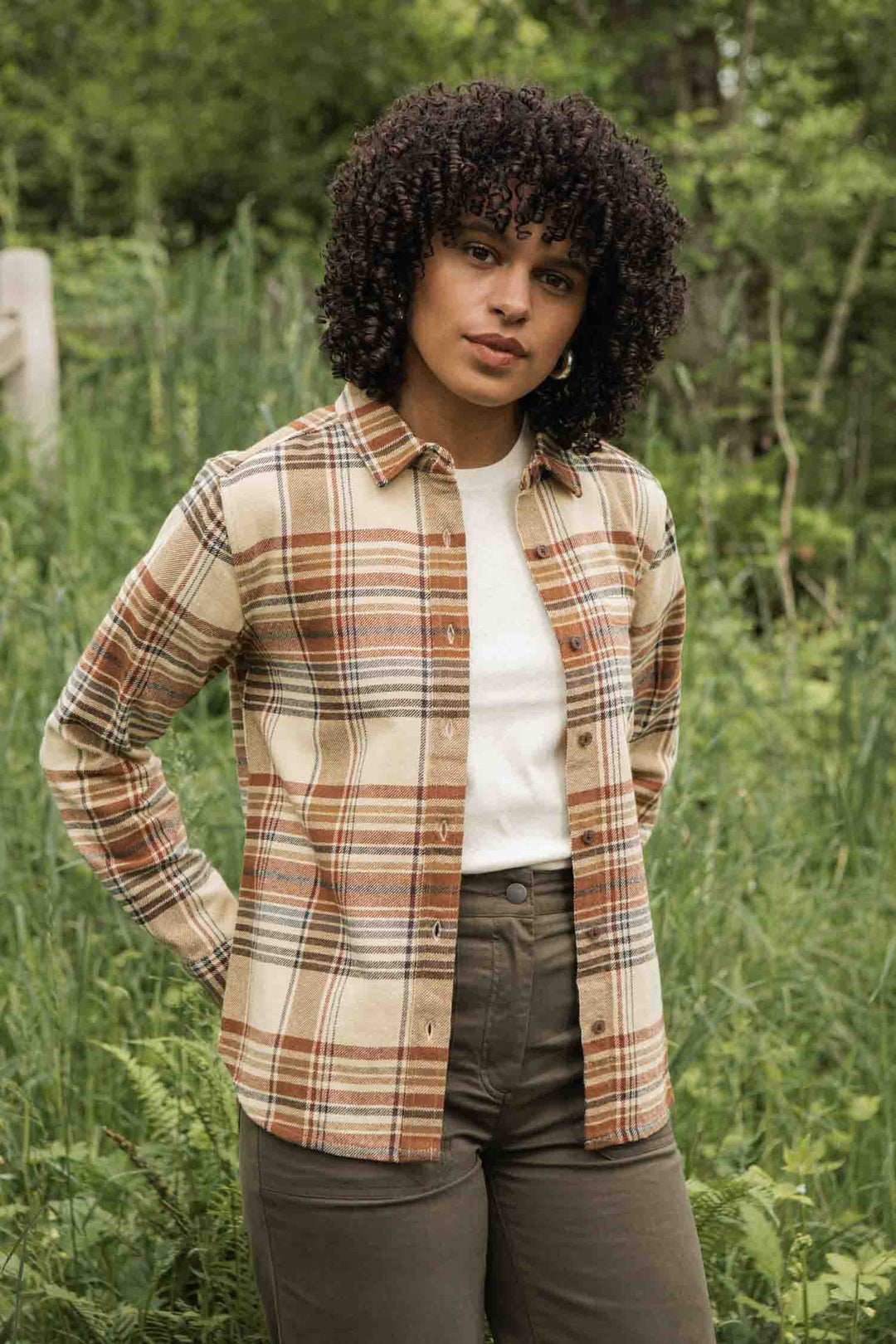 A person with curly hair stands in tall green grass, wearing the Bridge & Burn Ash Slim Shirt in Latte Plaid over a white top and olive green pants, looking at the camera with a neutral expression.