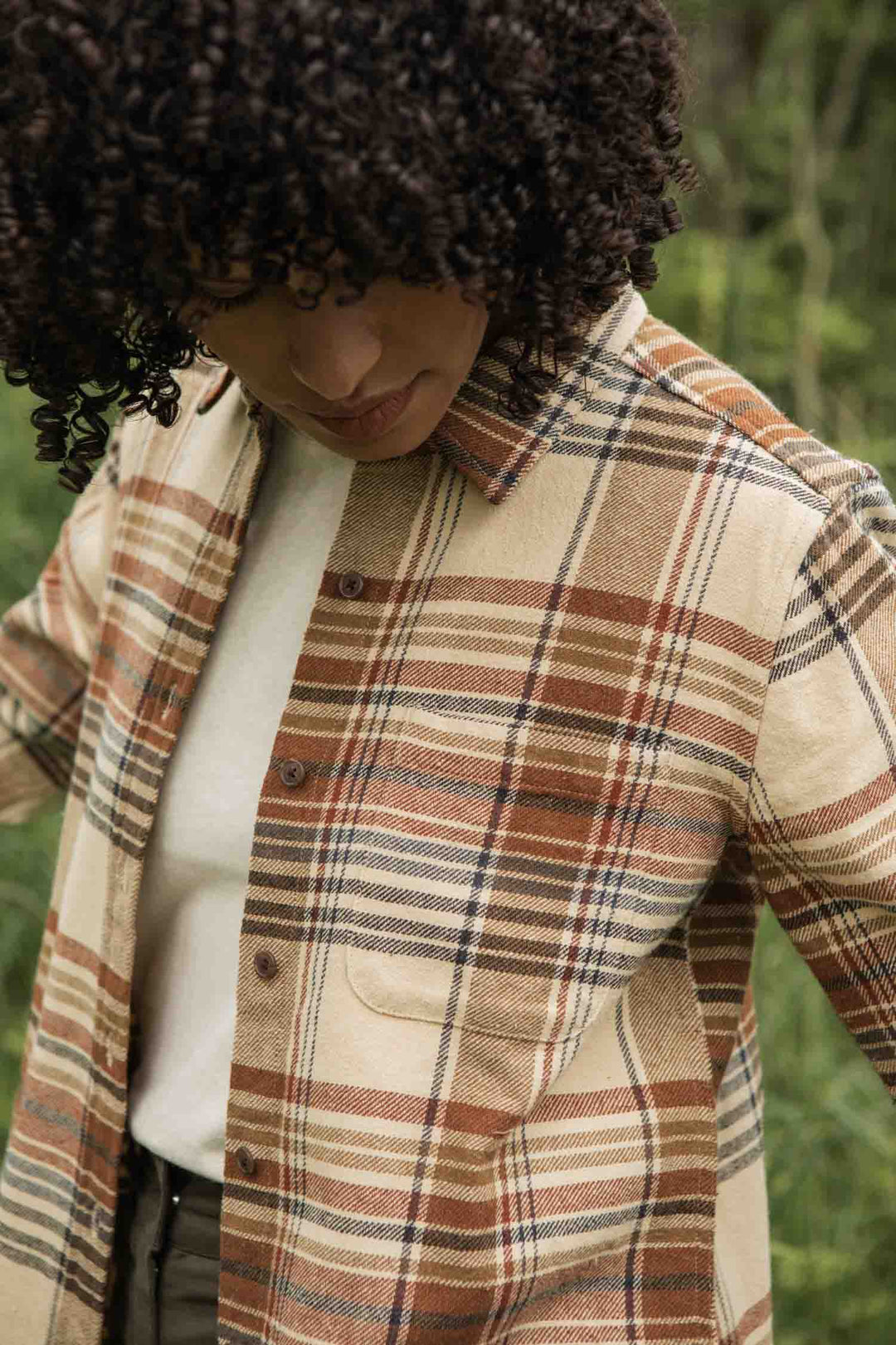A person with curly hair looks down while wearing the Bridge & Burn Ash Slim Shirt in Latte Plaid over a white t-shirt, standing outdoors with greenery in the background.
