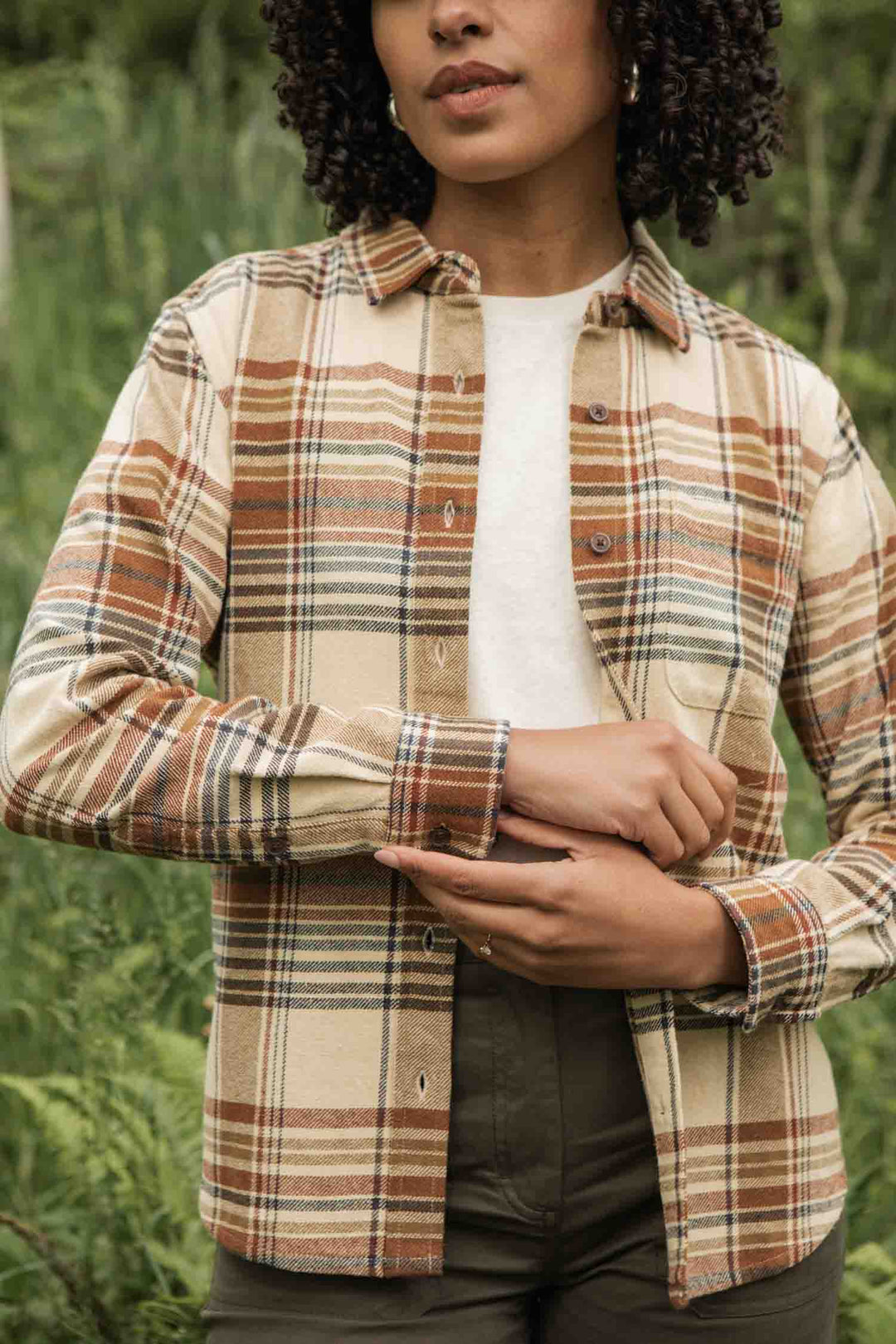 A person wears the Bridge & Burn Ash Slim Shirt in Latte Plaid—a slim fit button-up in beige, brown, and green—over a white top while standing outdoors with their hands clasped at the wrists.
