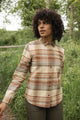 A person with curly hair wears the Bridge & Burn Ash Slim Shirt in Latte Plaid, standing outdoors on green grass with trees behind them, looking to the side.
