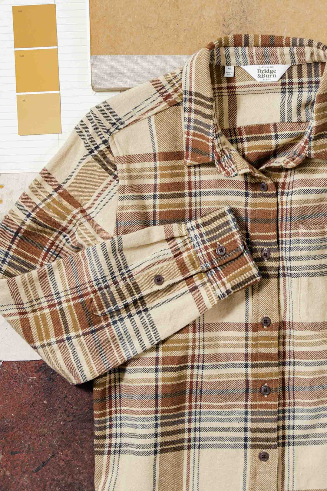The Bridge & Burn Ash Slim Shirt in Latte Plaid, featuring beige, brown, and rust tones, is laid flat beside neutral paint swatches and textured papers.