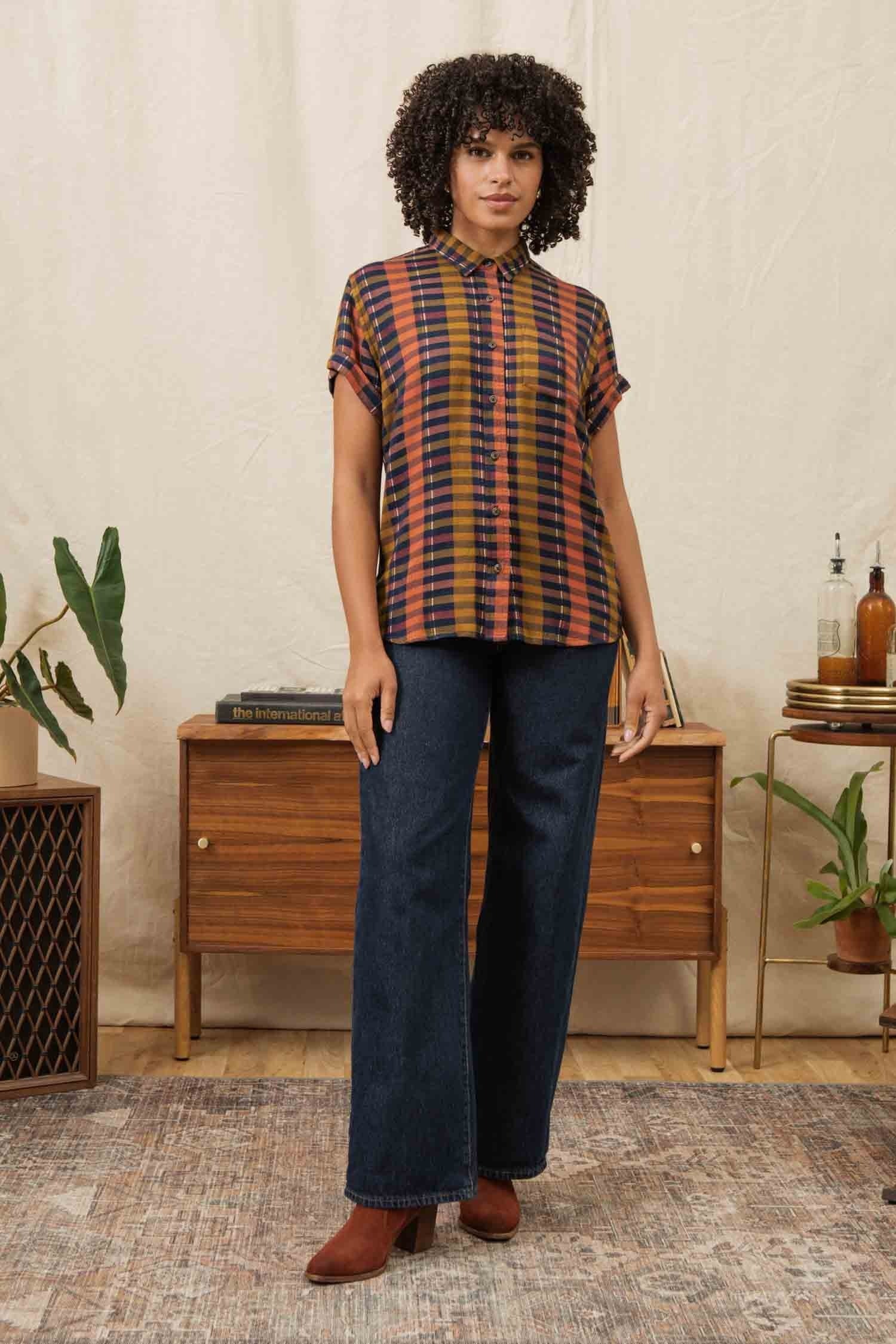 A woman with curly hair wears the Bridge & Burn Bea Shirt in Alpenglow Stripe, a tailored fit short-sleeve button down, paired with wide-leg blue jeans. She stands indoors on a patterned rug before a wooden sideboard with plants and decor.
