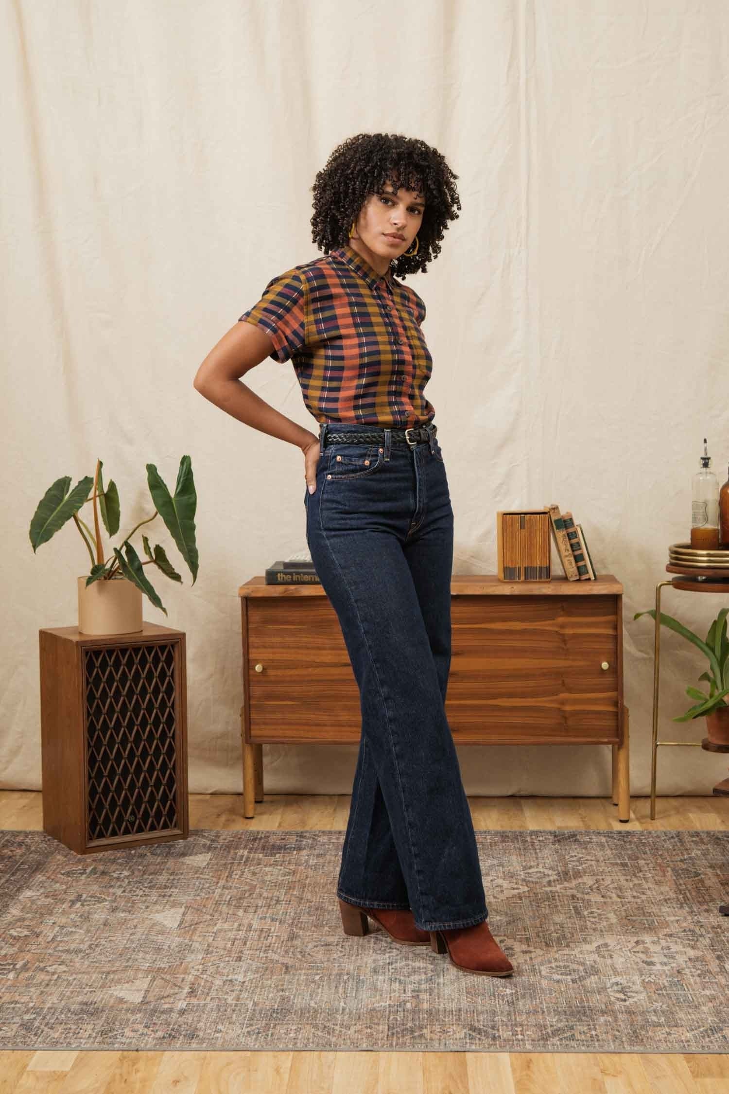 A woman with curly hair wears the Bridge & Burn Bea Shirt in Alpenglow Stripe and high-waisted dark blue jeans with brown boots. She stands indoors on a rug, surrounded by wooden furniture and plants in the background.