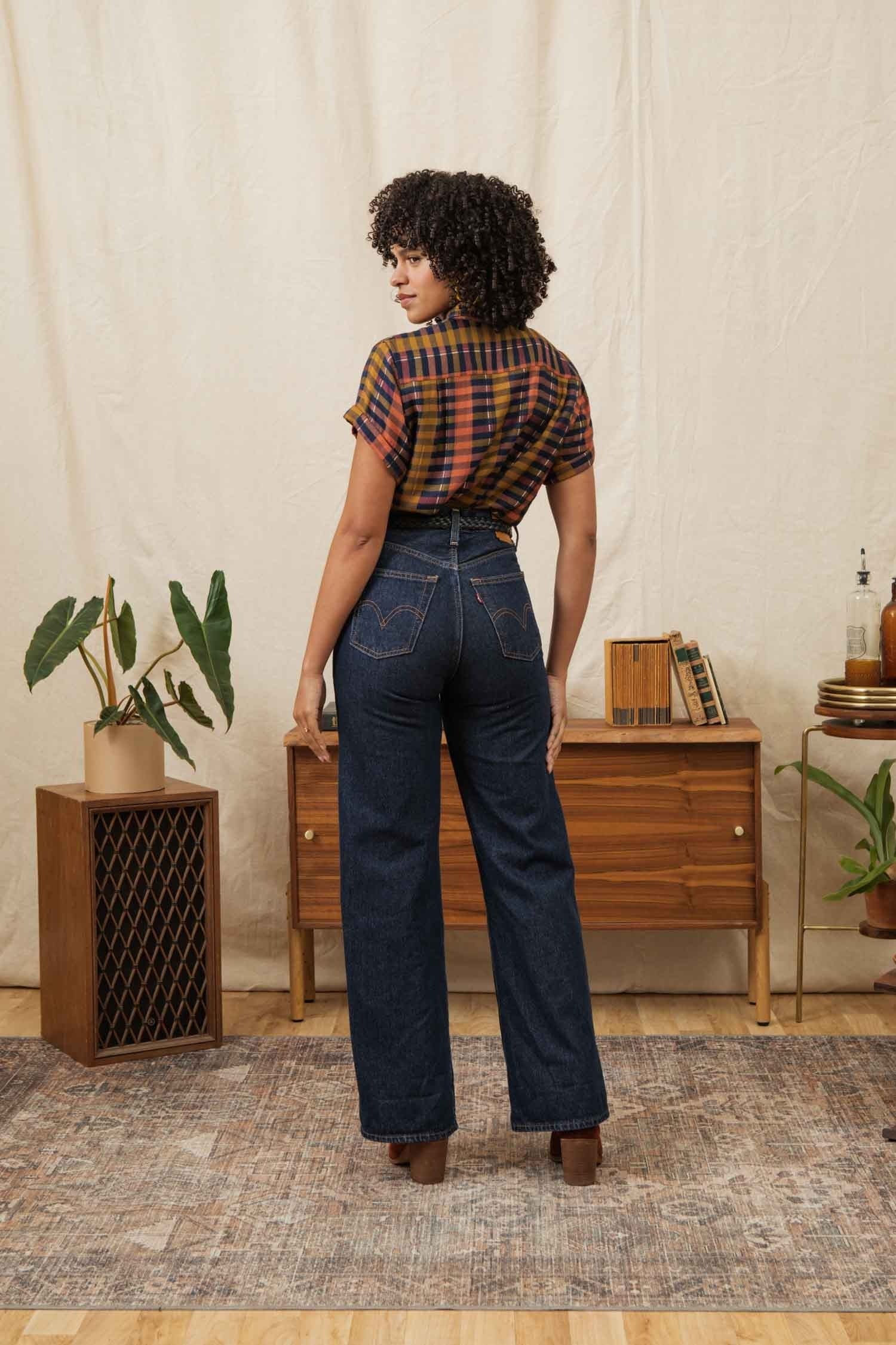 A woman with curly hair stands facing away, wearing the Bridge & Burn Bea Shirt in Alpenglow Stripe and high-waisted dark blue jeans. She is in a room with a wooden cabinet, plants, and décor on a beige rug and background.