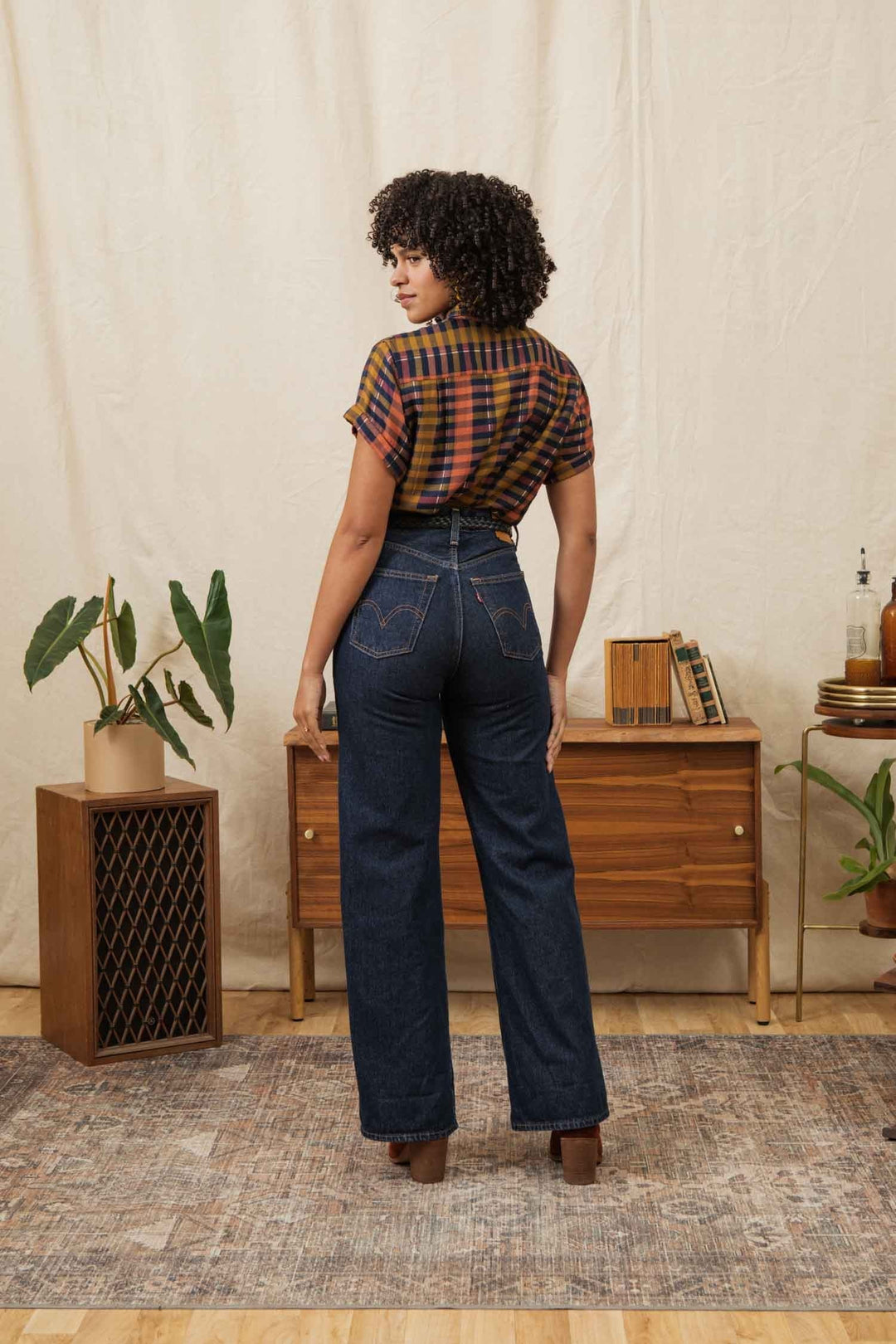 A woman with curly hair stands facing away, wearing the Bridge & Burn Bea Shirt in Alpenglow Stripe and high-waisted dark blue jeans. She is in a room with a wooden cabinet, plants, and décor on a beige rug and background.