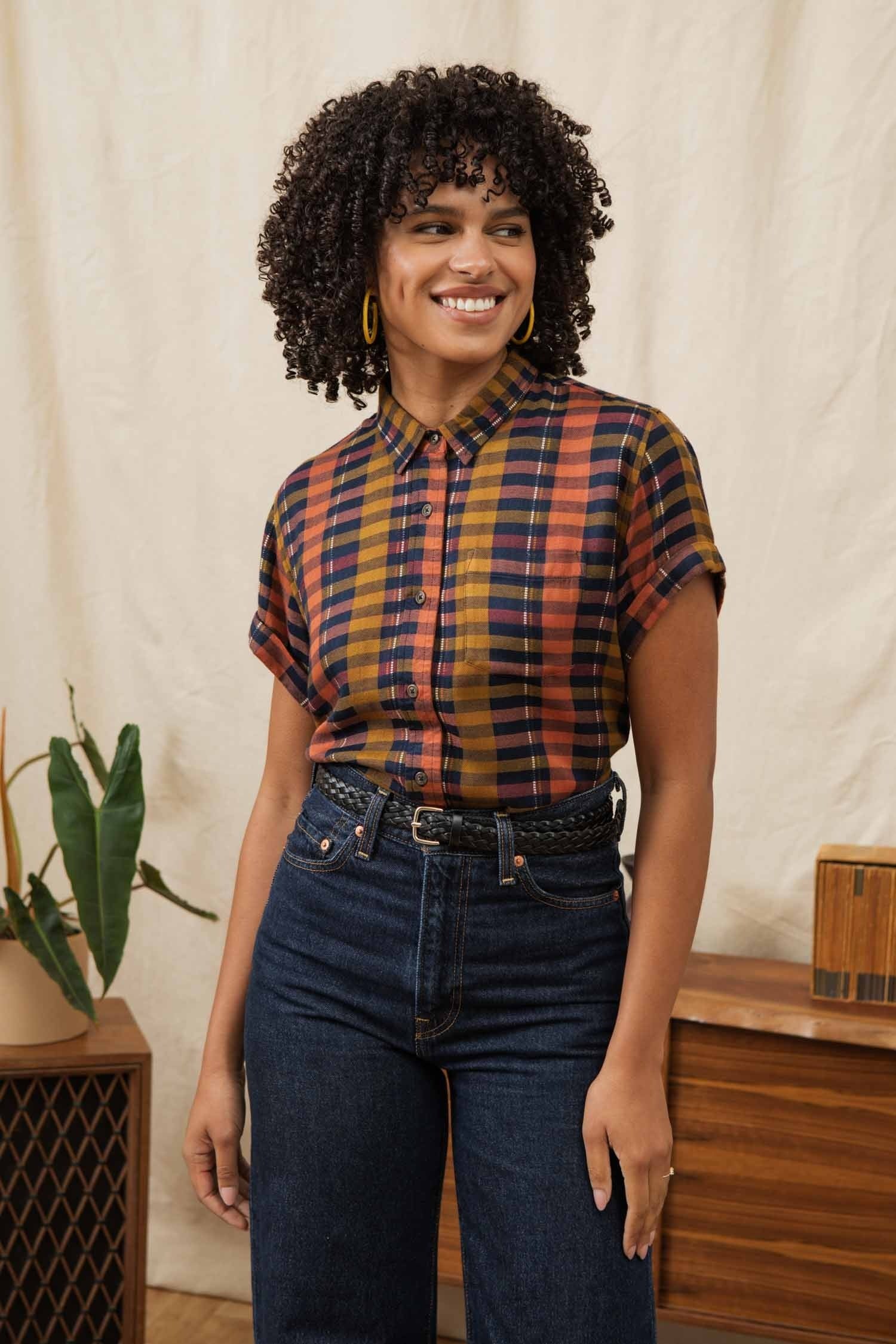 A woman with curly hair smiles indoors, wearing the Bridge & Burn Bea Shirt in Alpenglow Stripe and high-waisted dark jeans. A plant and wooden furniture can be seen in the background.