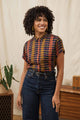 A woman with curly hair smiles indoors, wearing the Bridge & Burn Bea Shirt in Alpenglow Stripe and high-waisted dark jeans. A plant and wooden furniture can be seen in the background.