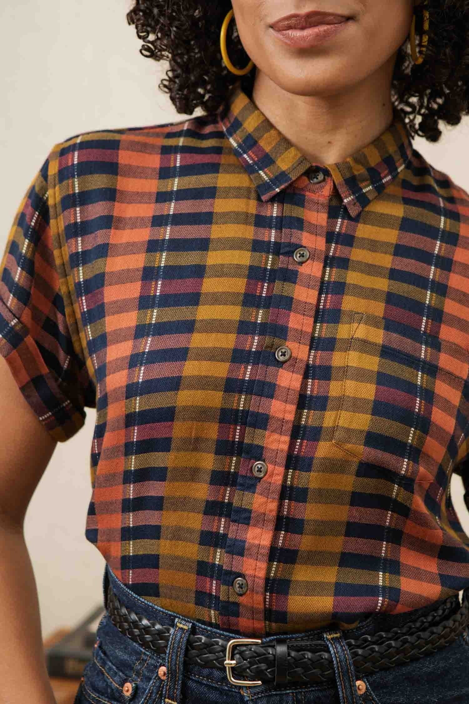 A woman wears the Bridge & Burn Bea Shirt in Alpenglow Stripe, a tailored fit short-sleeve button-down with orange, navy, and mustard stripes, tucked into high-waisted dark jeans and styled with a braided black belt.