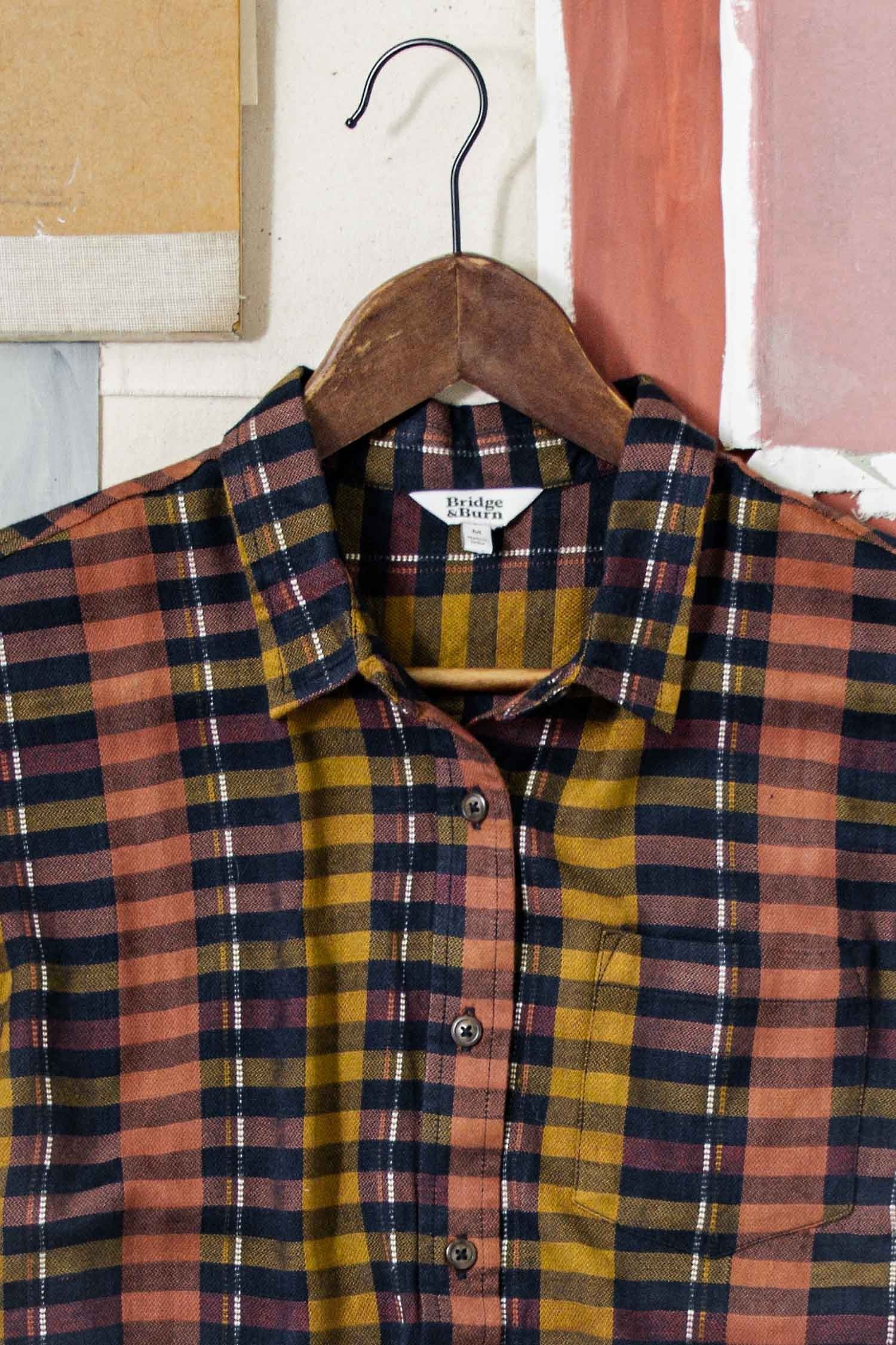 The Bridge & Burn Bea Shirt in Alpenglow Stripe, an eco-friendly short-sleeve button-down, features mustard yellow, rust, and black stripes and hangs on a wooden hanger against neutral and pinkish panels.