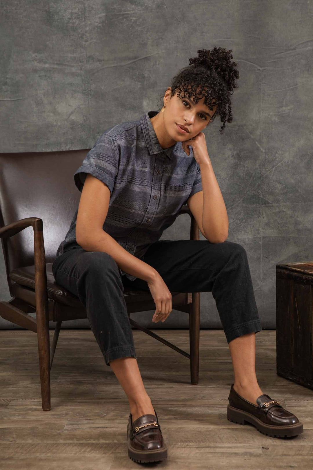 A person with curly hair sits on a brown chair, leaning their head on one hand. They wear the Bridge & Burn Bea Shirt in Cascade Weave, paired with dark pants and loafers. The backdrop is gray and neutral.