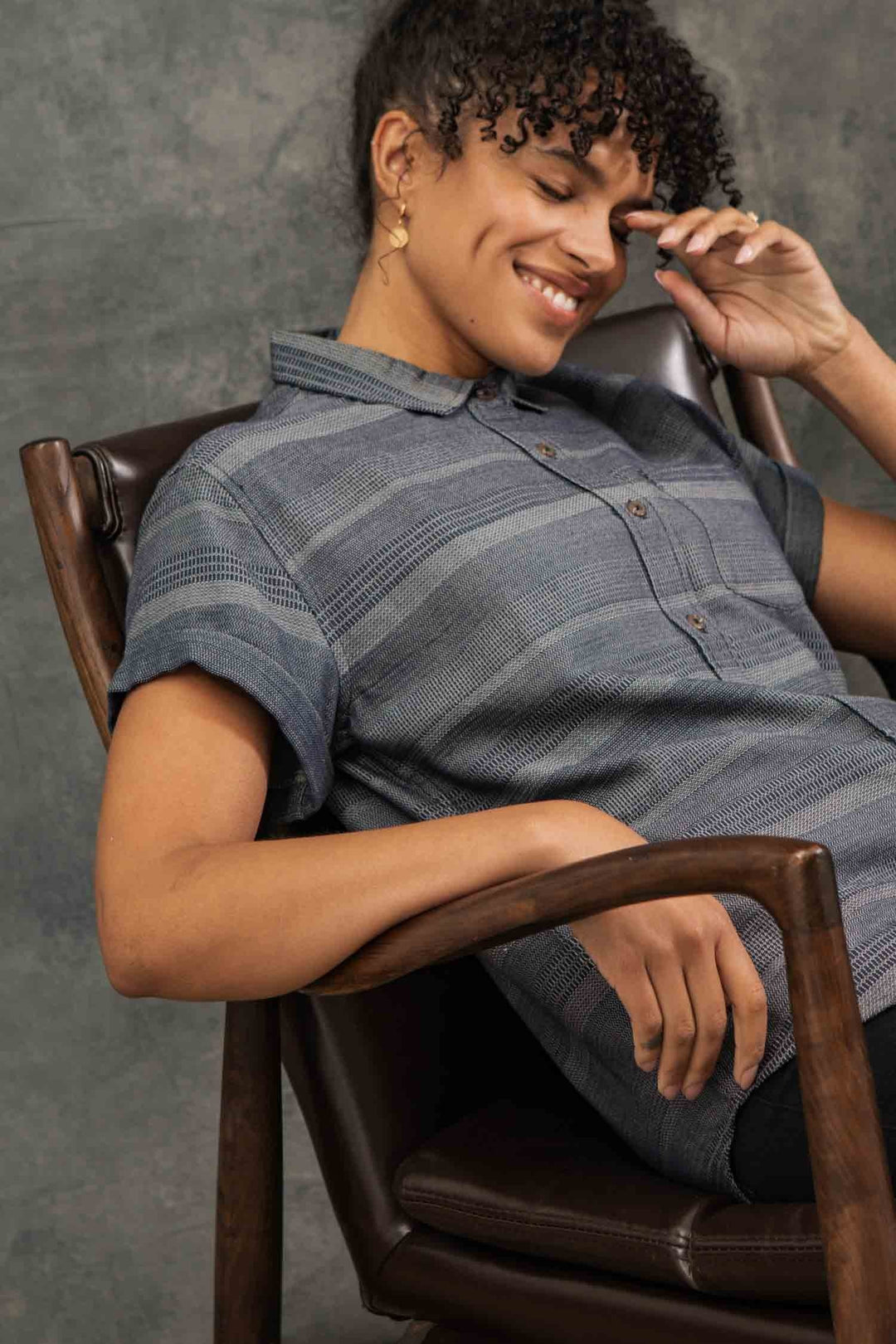 A person with curly hair sits in a brown leather chair, smiling with eyes closed, head resting on one hand. They wear the Bridge & Burn Bea Shirt in Cascade Weave and gold hoop earrings.