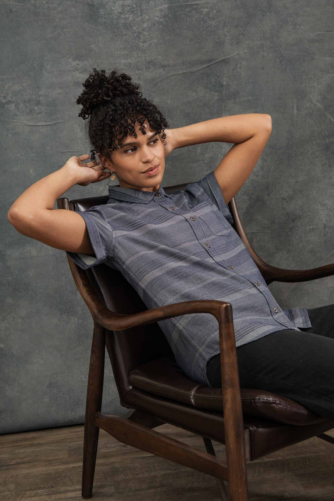 A person with curly hair leans back in a wooden chair, hands behind their head, wearing the Bridge & Burn Bea Shirt / Cascade Weave and dark pants, posed against a textured gray backdrop.