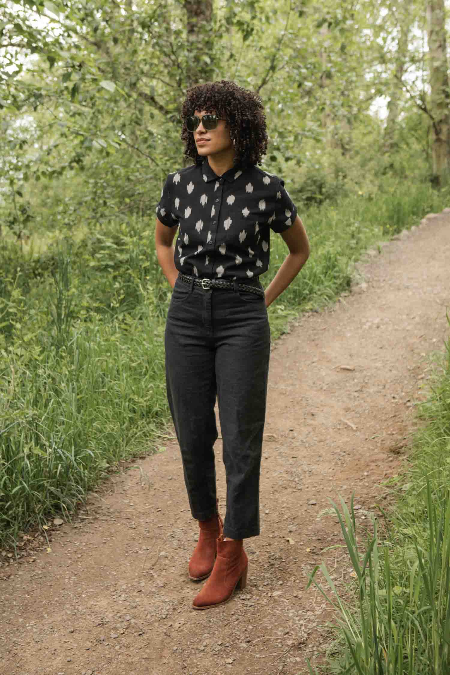A woman with curly hair and sunglasses walks on a park path, wearing the Bridge & Burn Bea Shirt in Eclipse Ikat, paired with high-waisted dark pants and red ankle boots.