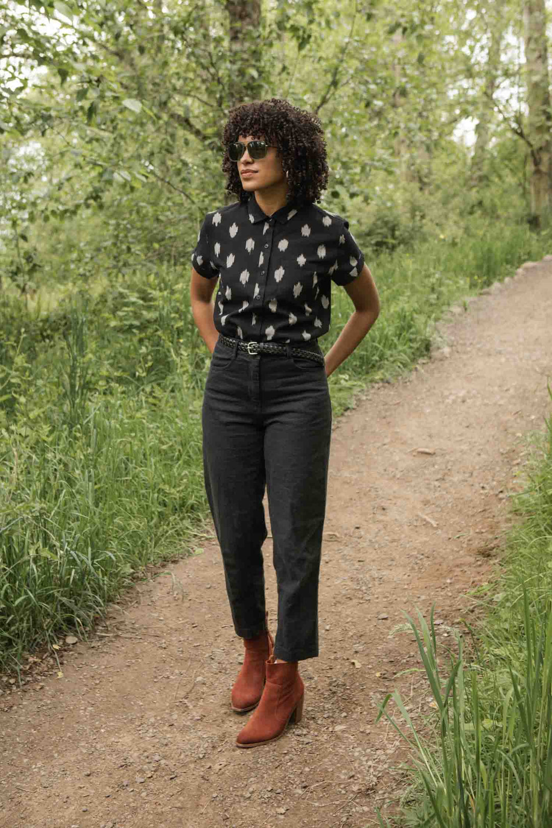A woman with curly hair and sunglasses walks on a park path, wearing the Bridge & Burn Bea Shirt in Eclipse Ikat, paired with high-waisted dark pants and red ankle boots.