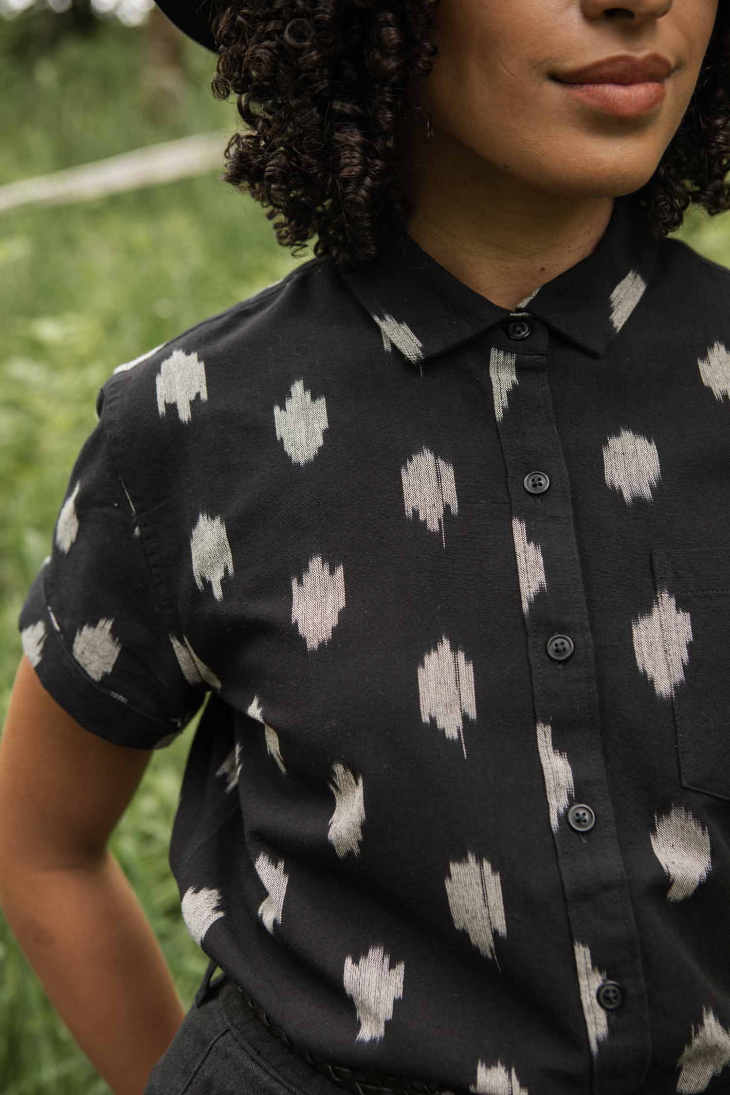 A person with curly hair stands calmly outdoors in a grassy area, wearing the Bridge & Burn Bea Shirt in Eclipse Ikat. Their face is partially visible.