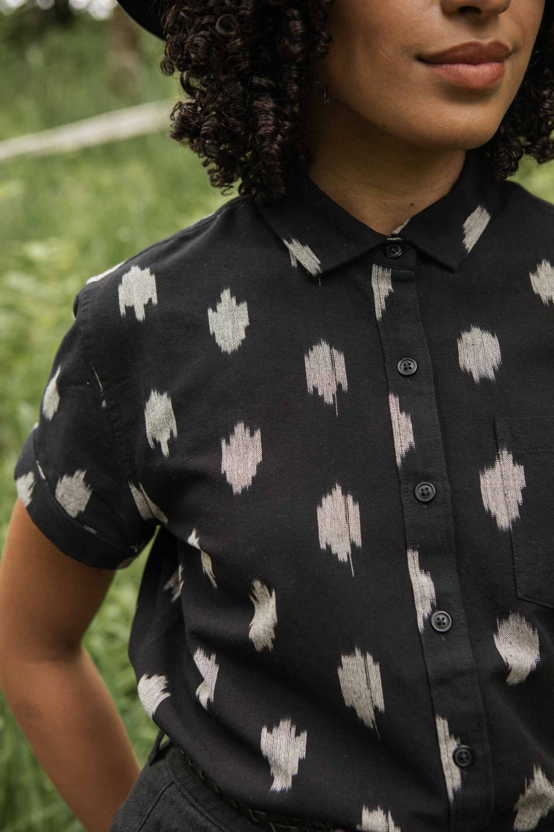 A person with curly hair stands calmly outdoors in a grassy area, wearing the Bridge & Burn Bea Shirt in Eclipse Ikat. Their face is partially visible.