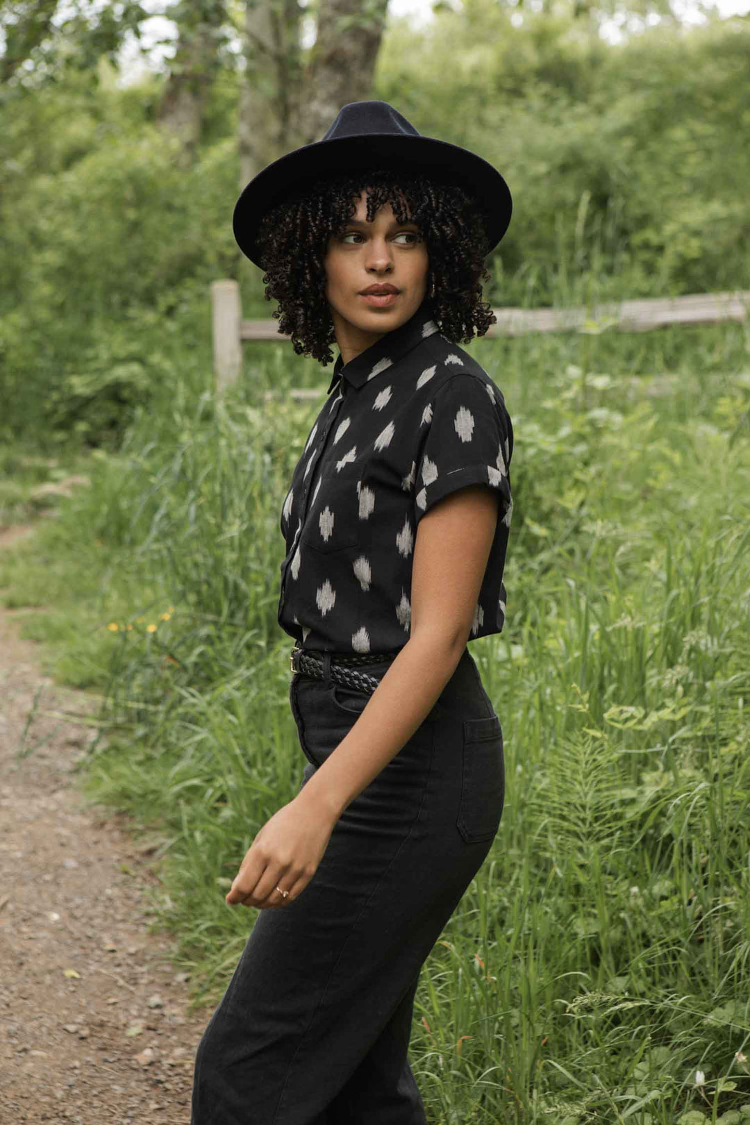 A woman with curly hair wears the Bridge & Burn Bea Shirt in Eclipse Ikat and high-waisted black pants, standing on a dirt path among green plants and trees, looking to the side.