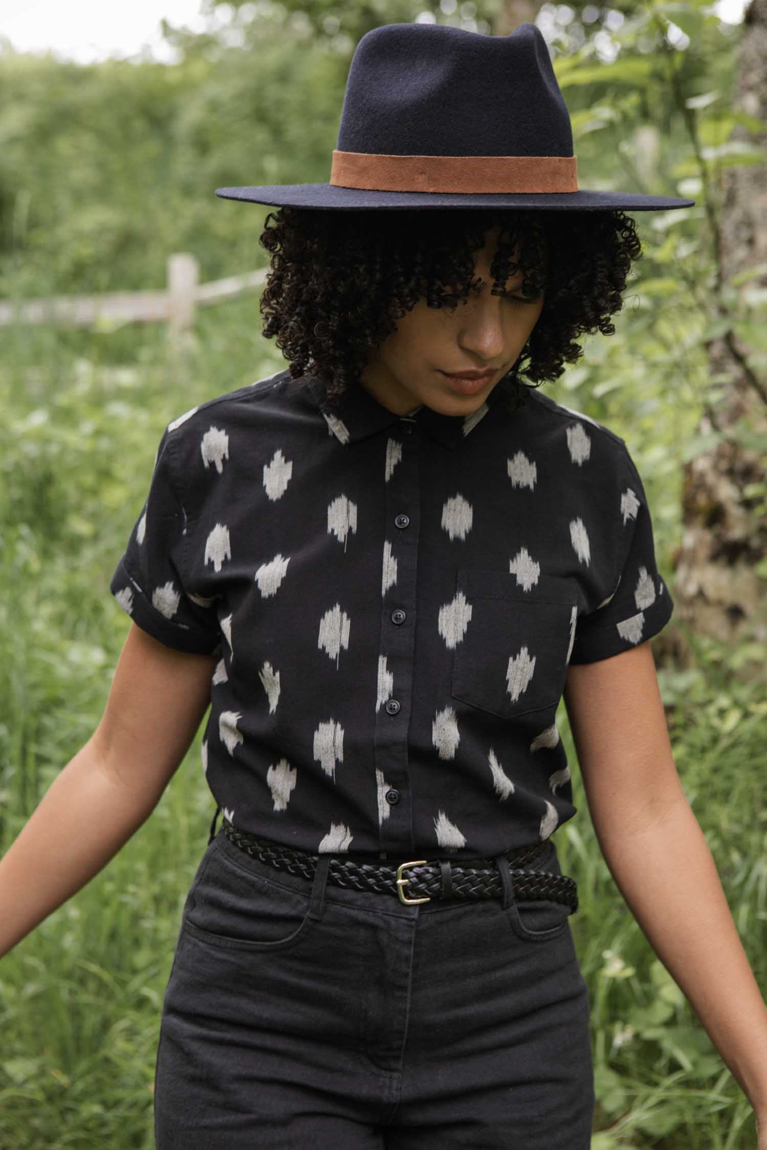 Wearing the Bridge & Burn Bea Shirt in Eclipse Ikat, paired with black pants and a wide-brimmed hat, a person stands outdoors among greenery, looking down with arms slightly out to the sides.