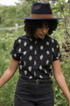 Wearing the Bridge & Burn Bea Shirt in Eclipse Ikat, paired with black pants and a wide-brimmed hat, a person stands outdoors among greenery, looking down with arms slightly out to the sides.