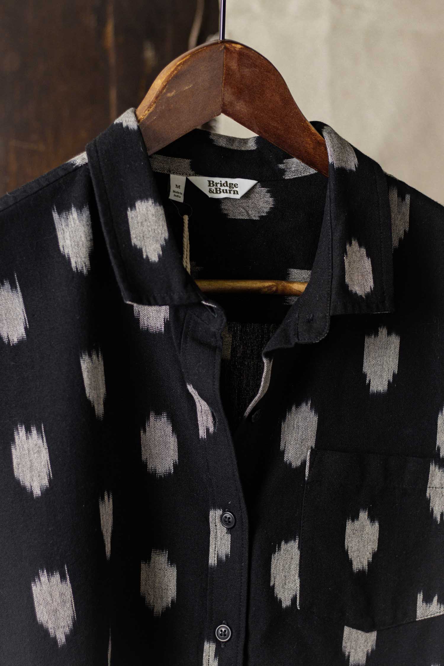 The Bea Shirt / Eclipse Ikat by Bridge & Burn is a black short-sleeve button down with a white dot pattern, left chest pocket, and hangs on a wooden hanger.