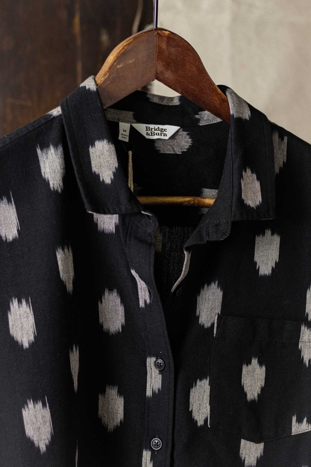 The Bea Shirt / Eclipse Ikat by Bridge & Burn is a black short-sleeve button down with a white dot pattern, left chest pocket, and hangs on a wooden hanger.