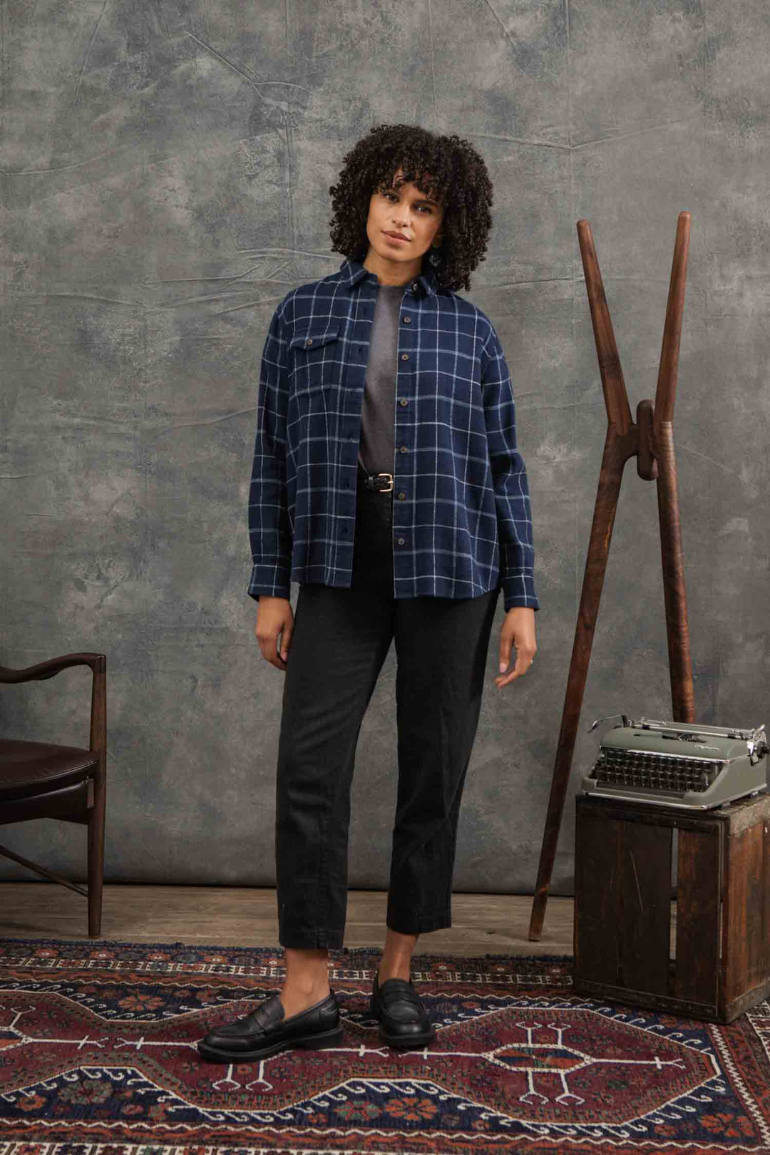 A woman with curly hair stands on a patterned rug, wearing the Bridge & Burn Bird Shirt in Moonrise Windowpane over a dark top, black pants, and black shoes. A vintage typewriter and wooden easel are beside her against a gray textured backdrop.