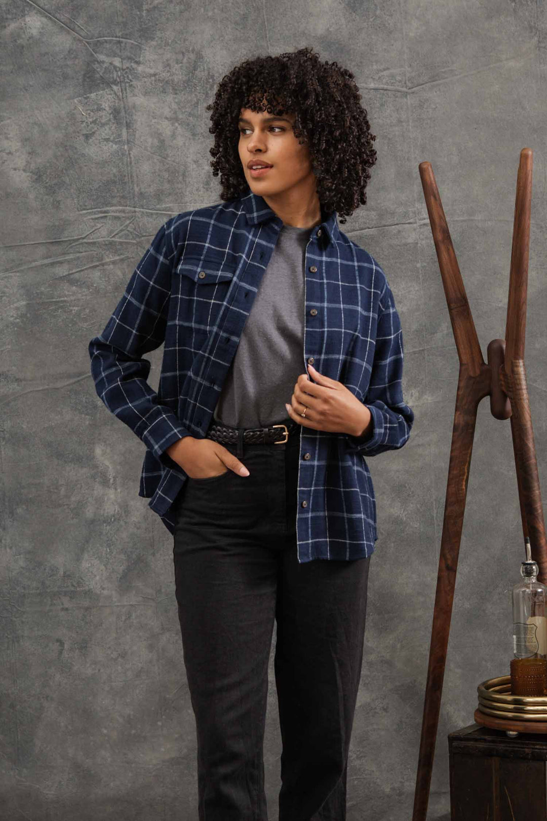 A person with curly hair wears the relaxed fit Bridge & Burn Bird Shirt in Moonrise Windowpane over a gray t-shirt and black pants, standing indoors near a wooden coat rack, small table with plates and bottle, against a textured gray backdrop.