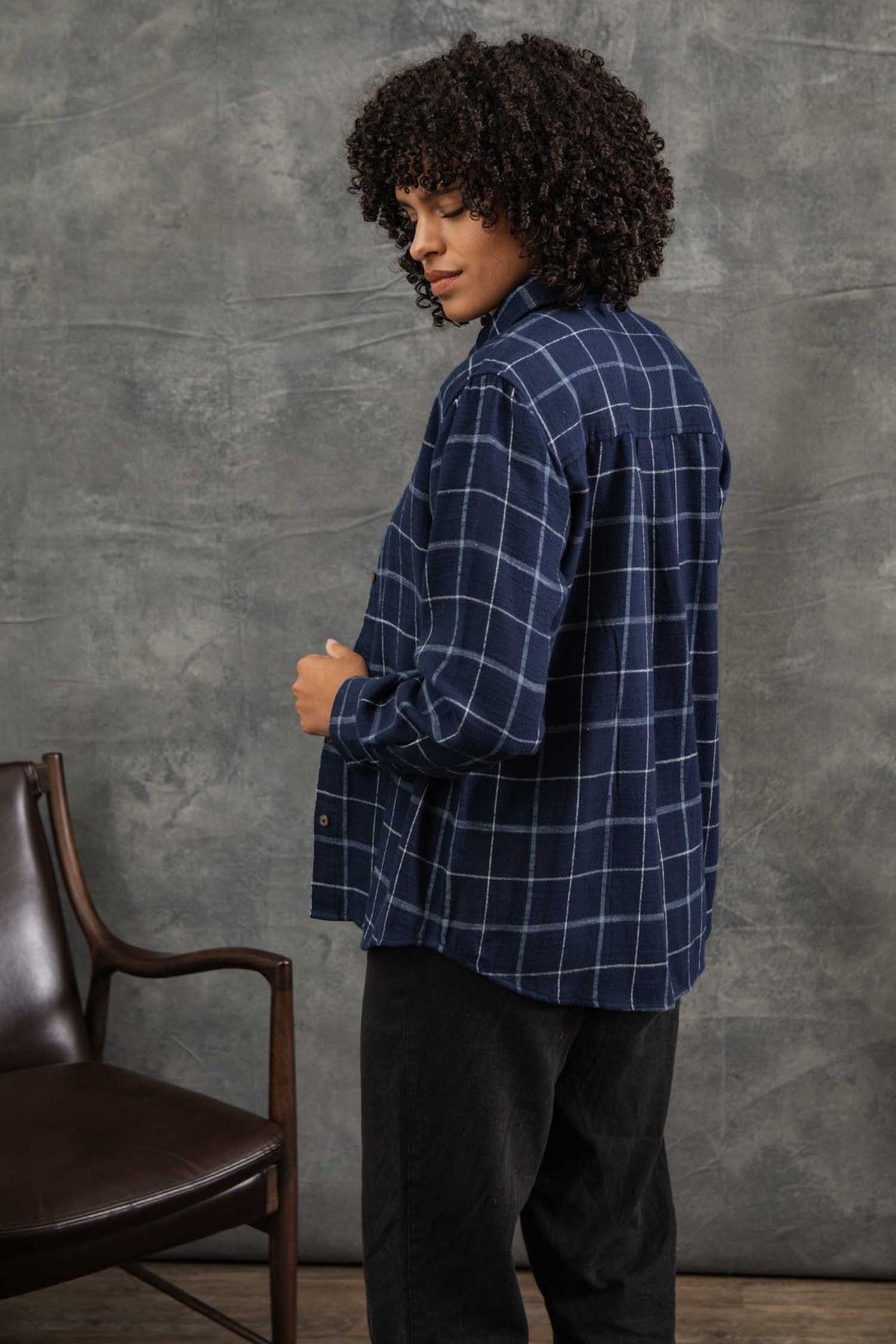 A person with curly hair wears a relaxed fit Bird Shirt / Moonrise Windowpane by Bridge & Burn and black pants, standing sideways and looking over their shoulder. A brown chair sits to the left against a gray textured background.