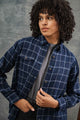 A person with curly hair wears the Bridge & Burn Bird Shirt in Moonrise Windowpane over a gray top, looking slightly to the side while adjusting the shirt against a neutral gray background.