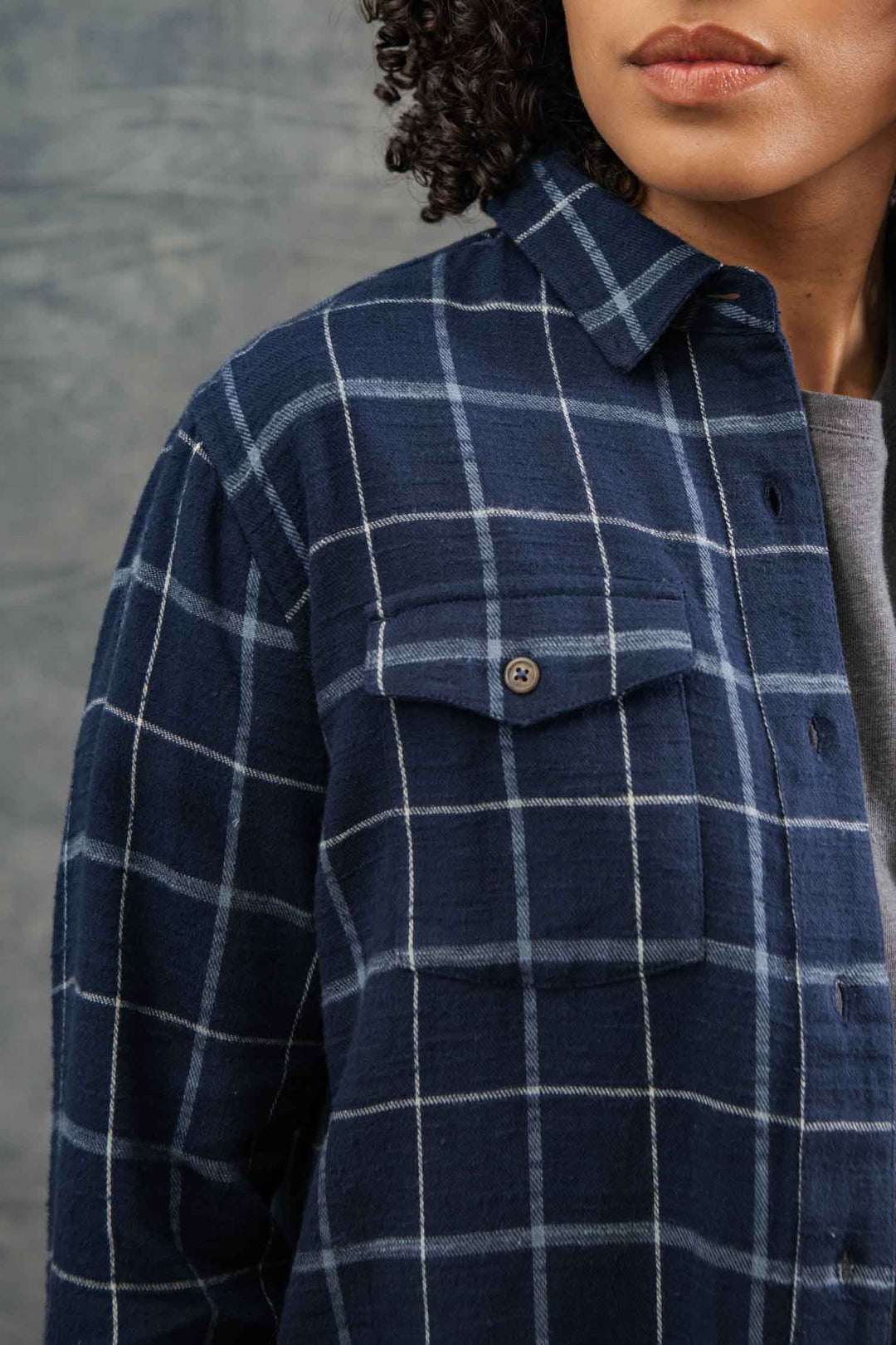 A person with curly hair wears the Bridge & Burn Bird Shirt in Moonrise Windowpane, a relaxed-fit blue and white plaid top with a front pocket, standing against a gray textured background. Only the lower face and upper body are visible.
