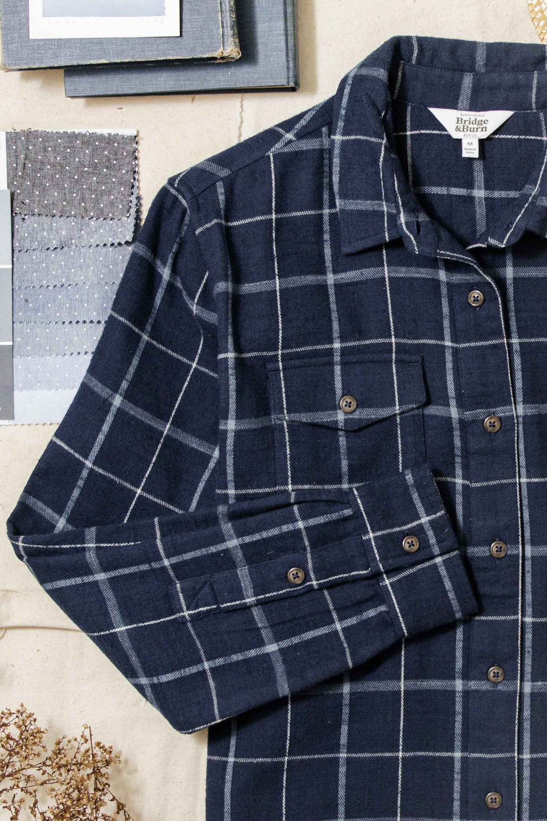 The Bird Shirt / Moonrise Windowpane by Bridge & Burn, a dark blue organic cotton flannel with a white windowpane plaid and relaxed fit, is displayed on a light surface beside fabric swatches and closed books in the background.
