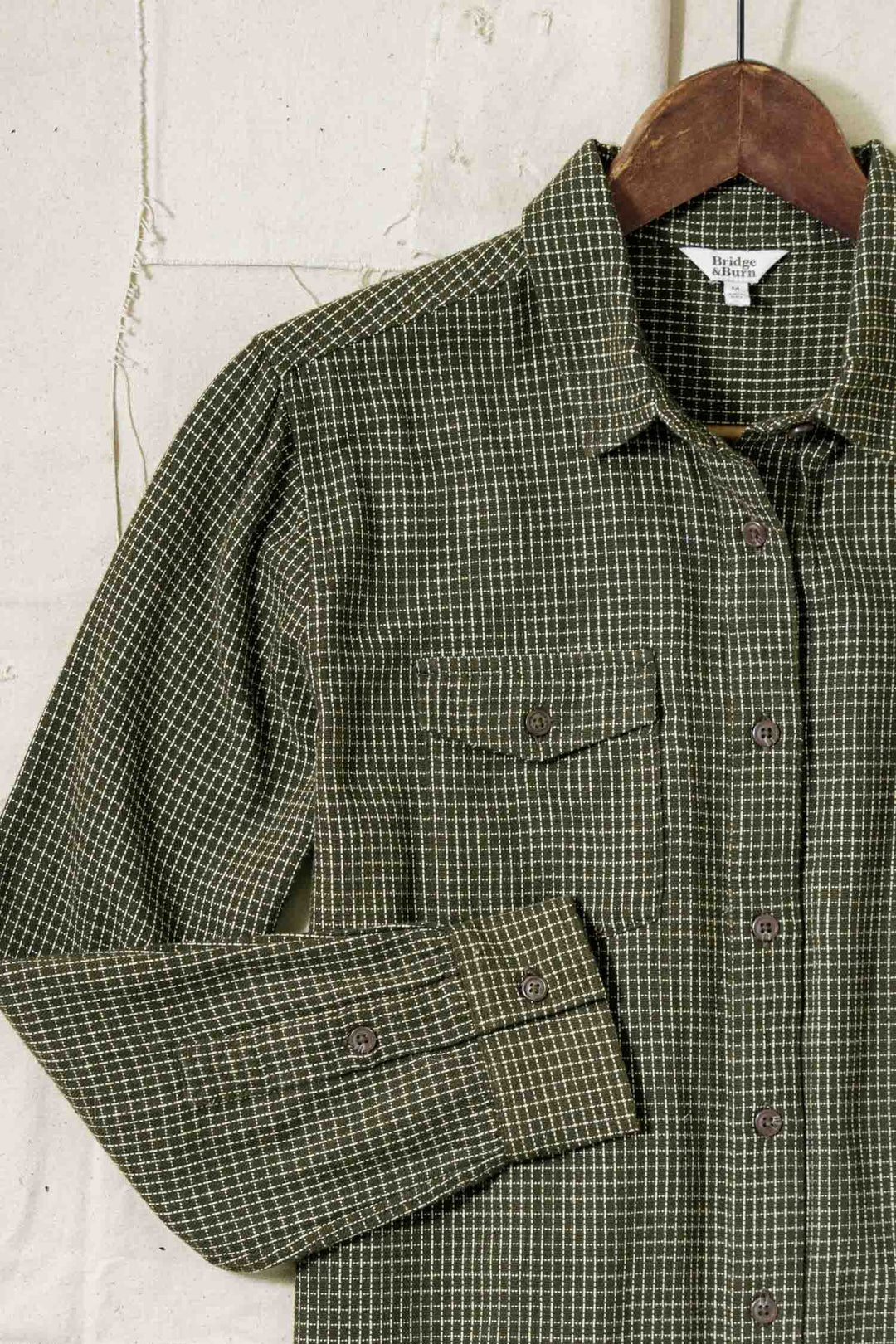 The Bird Shirt / Olive Grid Plaid by Bridge & Burn, a relaxed fit men's shirt with a chest pocket and visible stitching, hangs on a wooden hanger against a light textured wall. The green and white plaid design adds classic style.