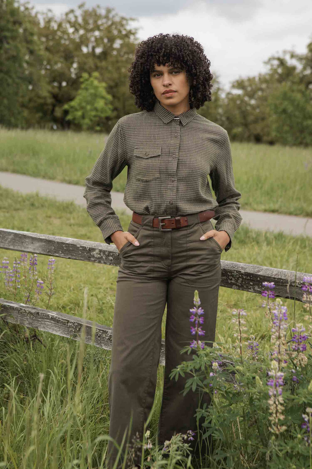 A person with curly hair stands outdoors by a wooden fence, wearing the Bridge & Burn Bird Shirt in Olive Grid Plaid tucked into high-waisted pants with a brown belt. Purple wildflowers, green grass, and trees complete the scenic backdrop.