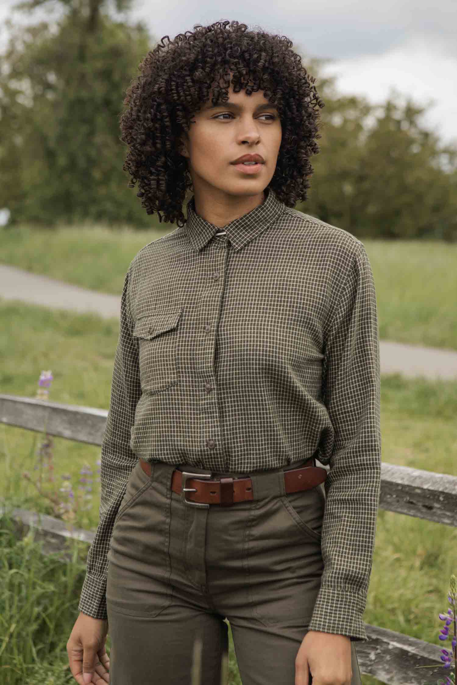 A person with curly hair wears the Bridge & Burn Bird Shirt in Olive Grid Plaid and green pants, standing outdoors before a wooden fence with greenery and a path in the background.