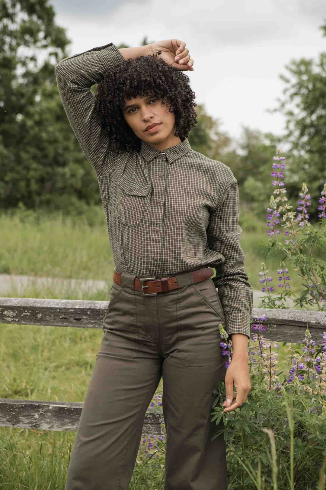 A curly-haired person stands outdoors by a wooden fence in green grass and purple wildflowers, wearing the Bridge & Burn Bird Shirt in Olive Grid Plaid with high-waisted pants. One arm is raised, resting on their head.