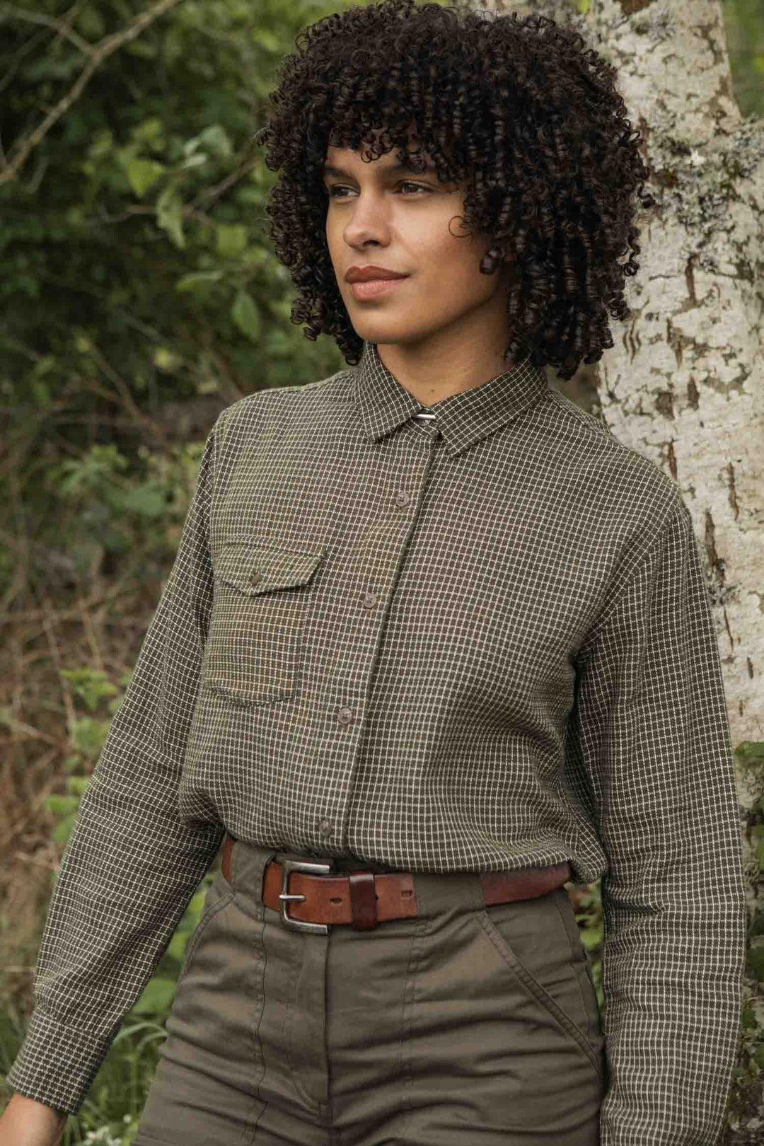 Wearing the Bridge & Burn Bird Shirt in Olive Grid Plaid, a person with curly hair and high-waisted trousers stands outside by a tree amid lush green foliage.