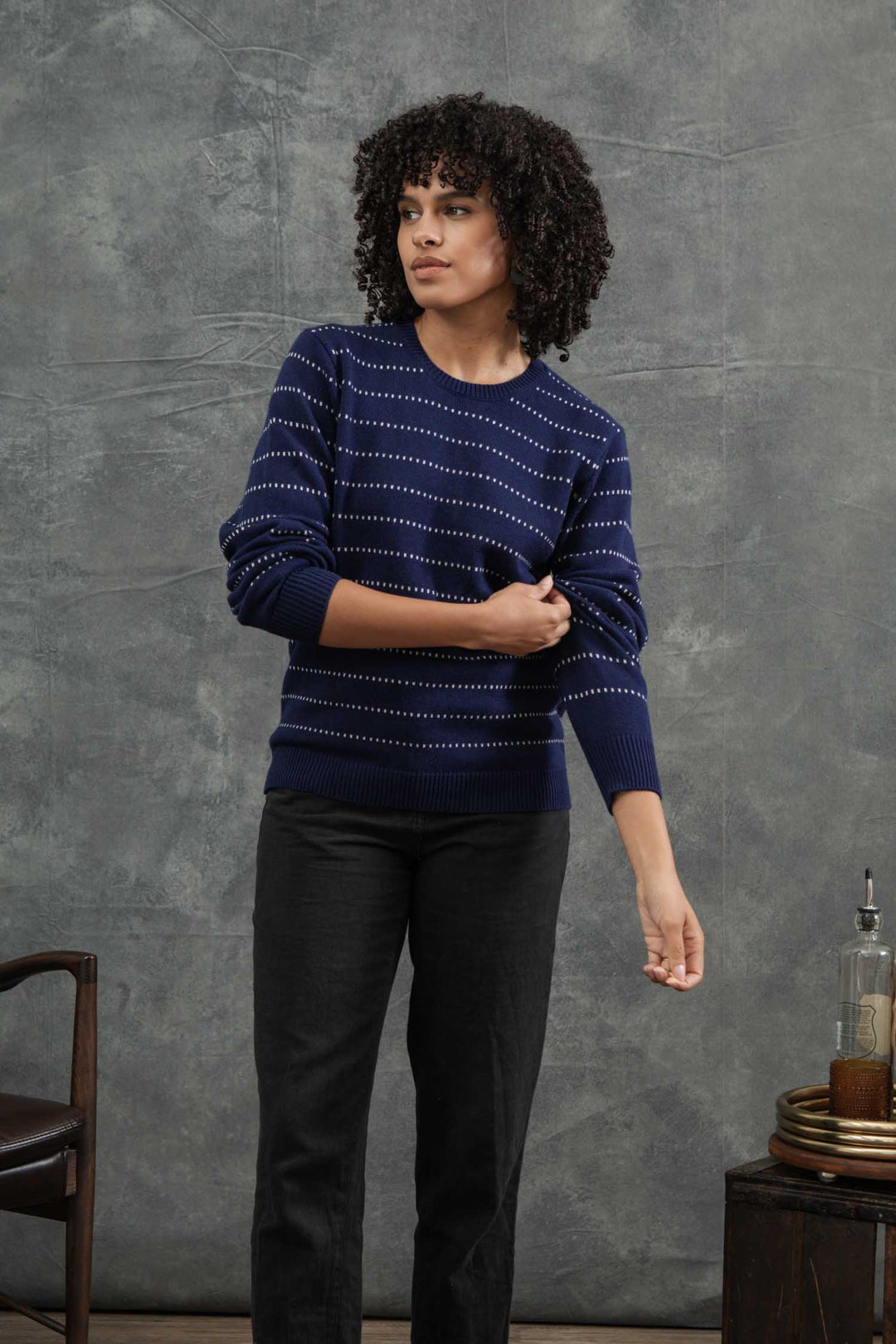A woman with curly hair wears the Bridge & Burn Cline Sweater in Navy Dotted Path and black pants, standing against a gray textured background as she adjusts her sleeve. A wooden chair and a small table are partially visible nearby.