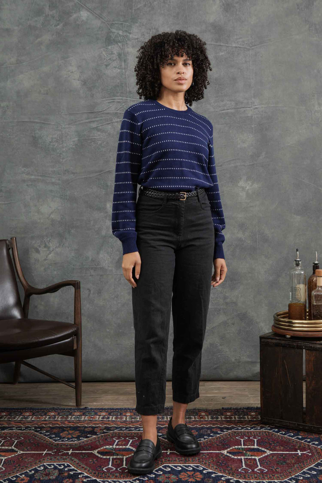 A woman with curly hair wears the Bridge & Burn Cline Sweater in Navy Dotted Path, paired with black high-waisted pants and black loafers, as she stands indoors on a patterned rug beside a wooden chair and a table with bottles.