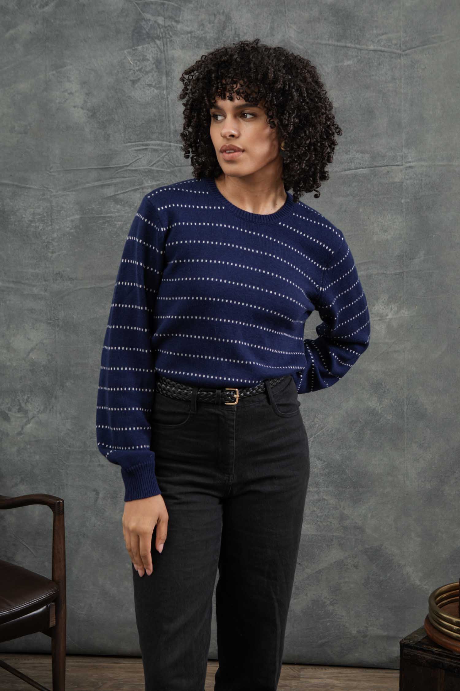 A woman with curly hair wears the Bridge & Burn Cline Sweater in Navy Dotted Path, paired with high-waisted black pants, standing before a gray backdrop next to a wooden chair.