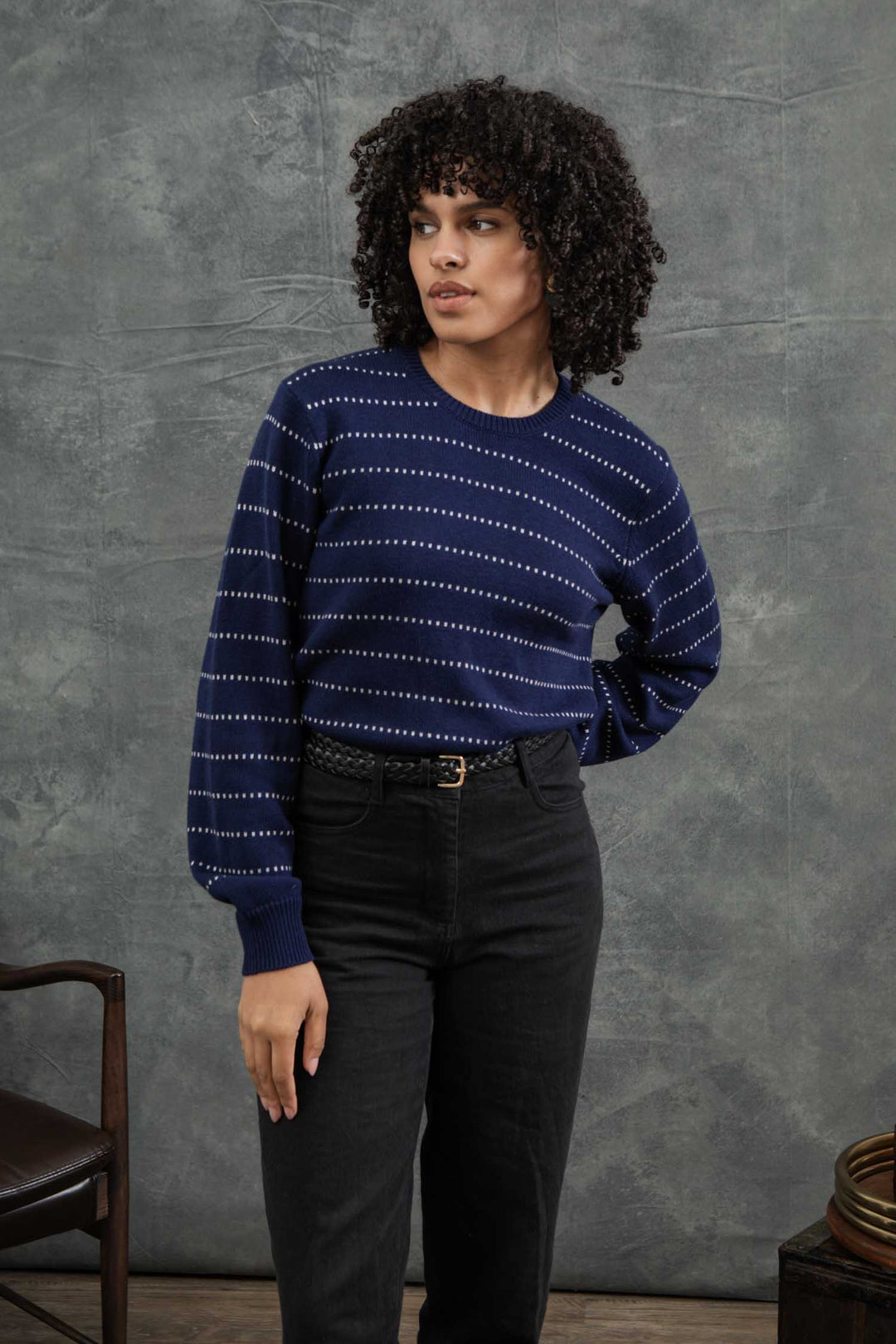 A woman with curly hair wears the Bridge & Burn Cline Sweater in Navy Dotted Path, paired with high-waisted black pants, standing before a gray backdrop next to a wooden chair.