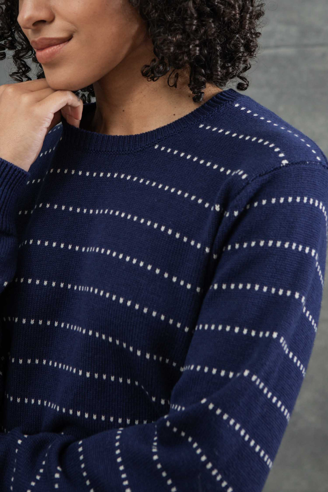 A person with curly hair models the Bridge & Burn Cline Sweater in Navy Dotted Path, featuring white dashed stripes, posing with one hand lightly touching their chin against a neutral background.