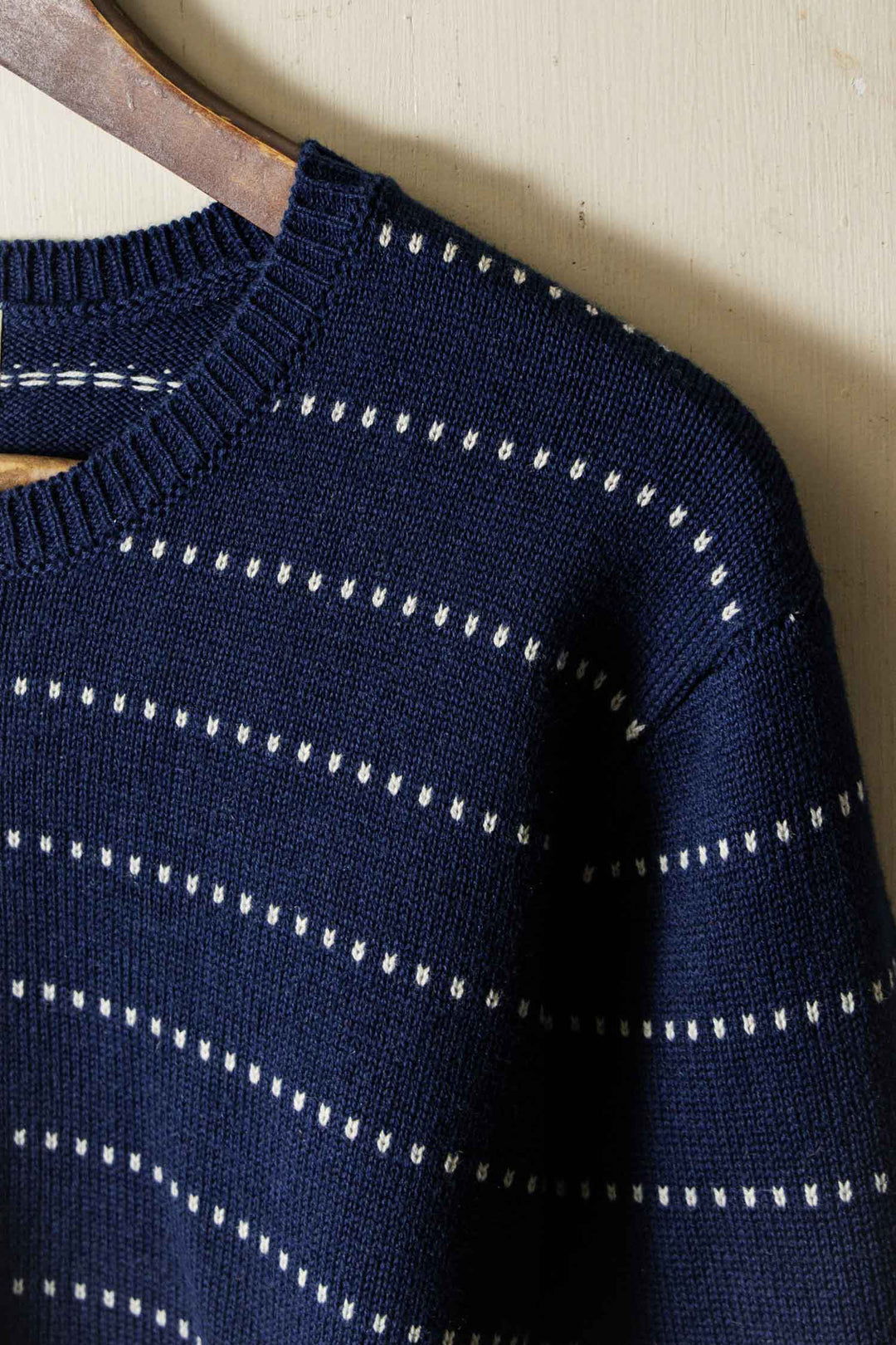 A close-up of the Bridge & Burn Cline Sweater in Navy Dotted Path—navy merino wool with small white horizontal dotted stripes—hanging on a wooden hanger against a light-colored wall.